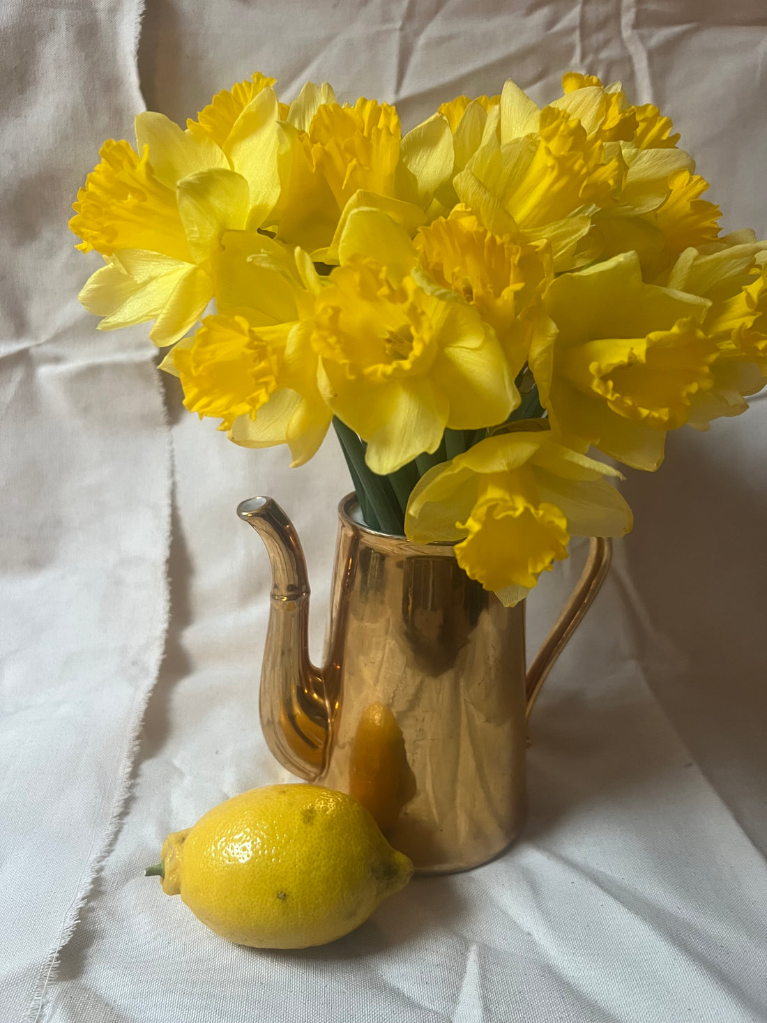 Yellow flowers in a gold vase with a lemon on a white fabric background